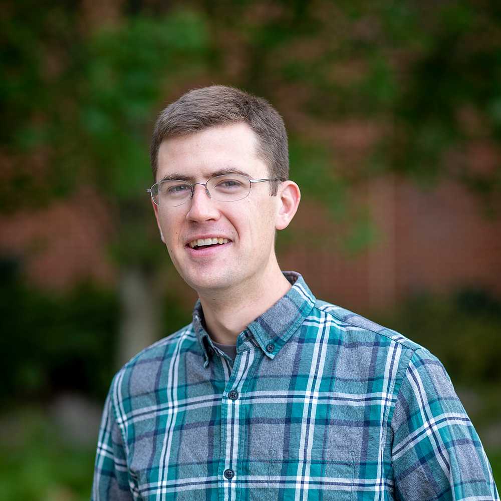 A photo of Christopher White, outdoors, wearing a flannel shirt, and smiling.